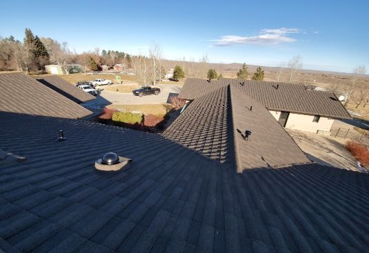 View from a shingled rooftop over suburban houses, trees, and open fields on a clear day