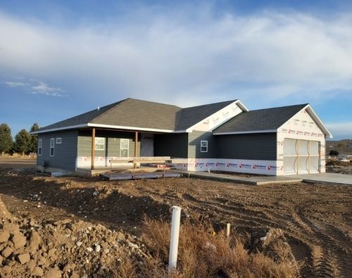 New house under construction with blue siding, dark roof, and dirt foreground under a cloudy sky