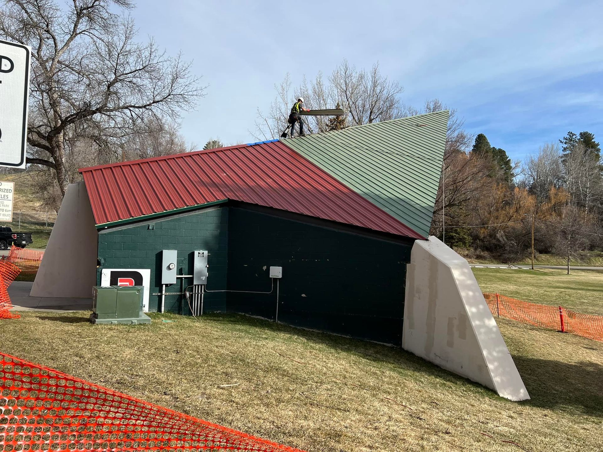 Small utility building with red and green roofs on grassy slope, beside orange fencing and trees.