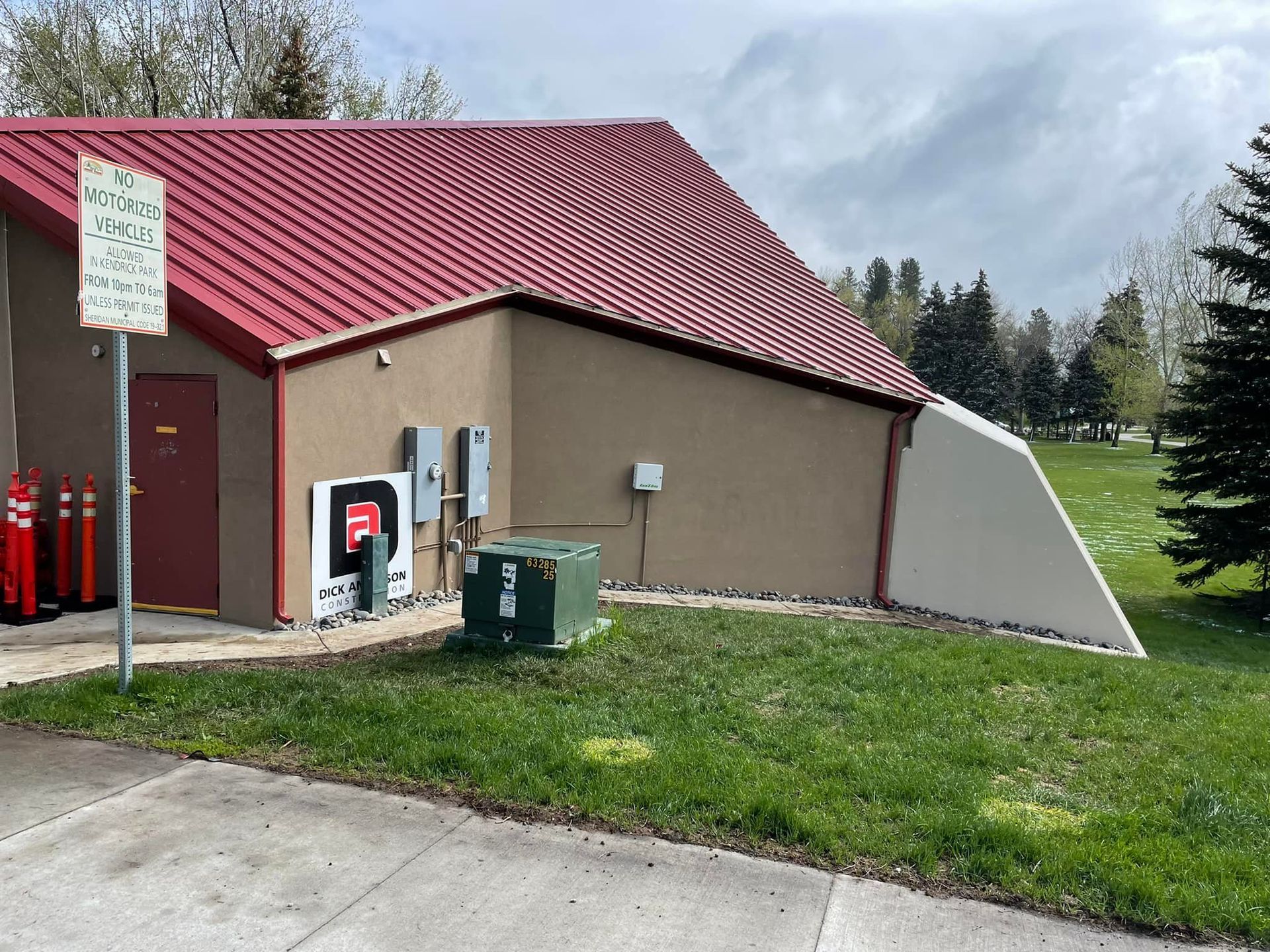 Small utility building with red roof beside a grassy roadside area and electrical equipment
