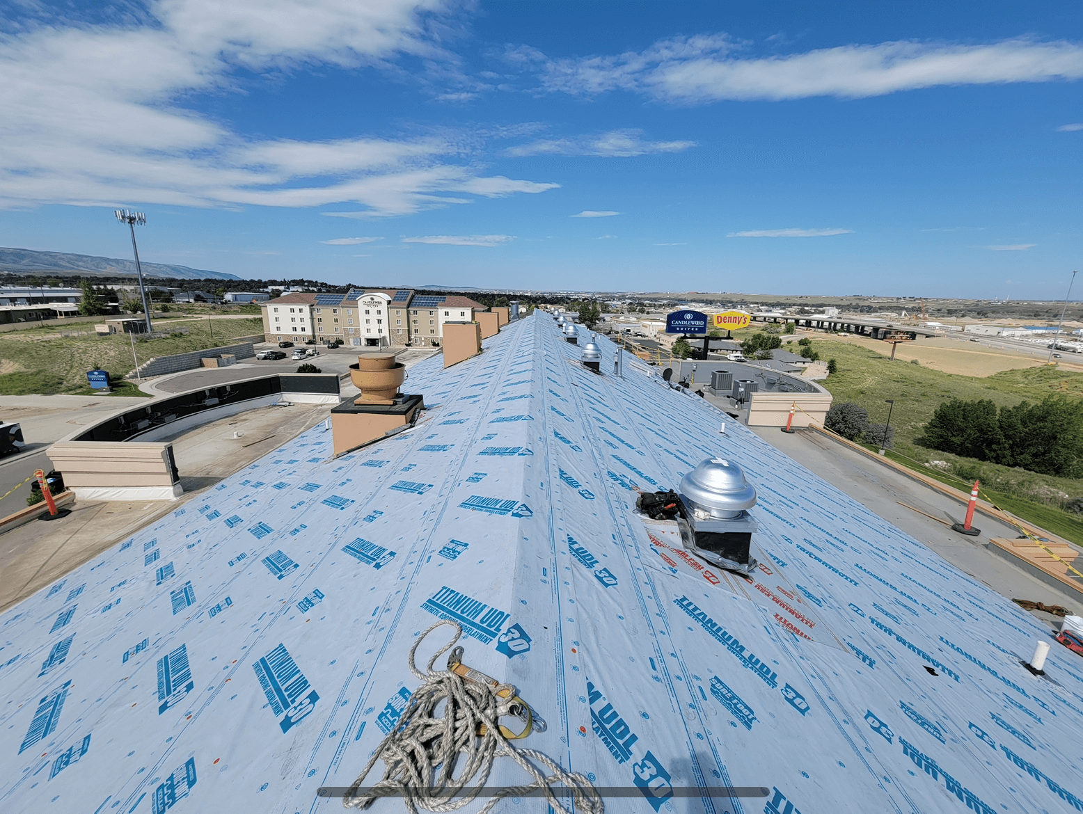 Rooftop under construction with blue protective wrap, vents, and a bright sky over a suburban area.
