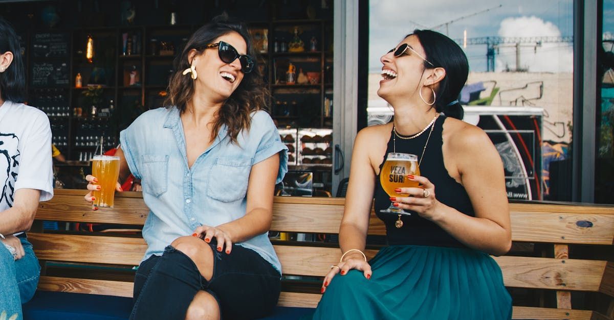 Two women are sitting on a bench laughing and drinking beer.