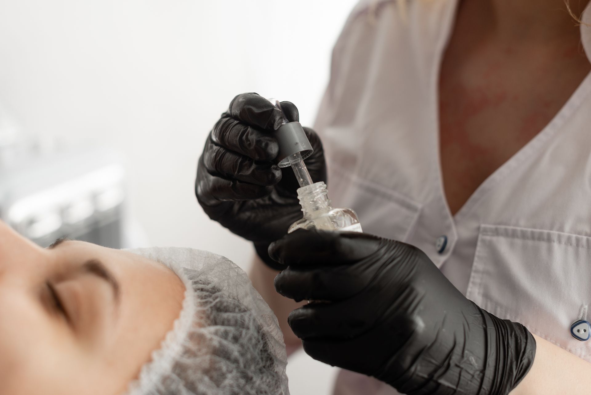 Technician in black gloves applying facial treatment to a client lying under a hair cap
