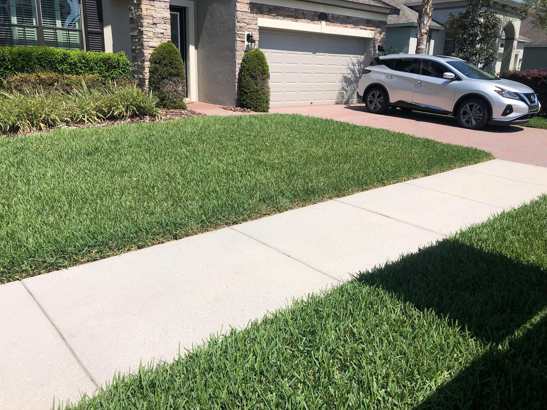 A suburban home with a green lawn, a concrete sidewalk in the foreground, and a silver car parked in the driveway.