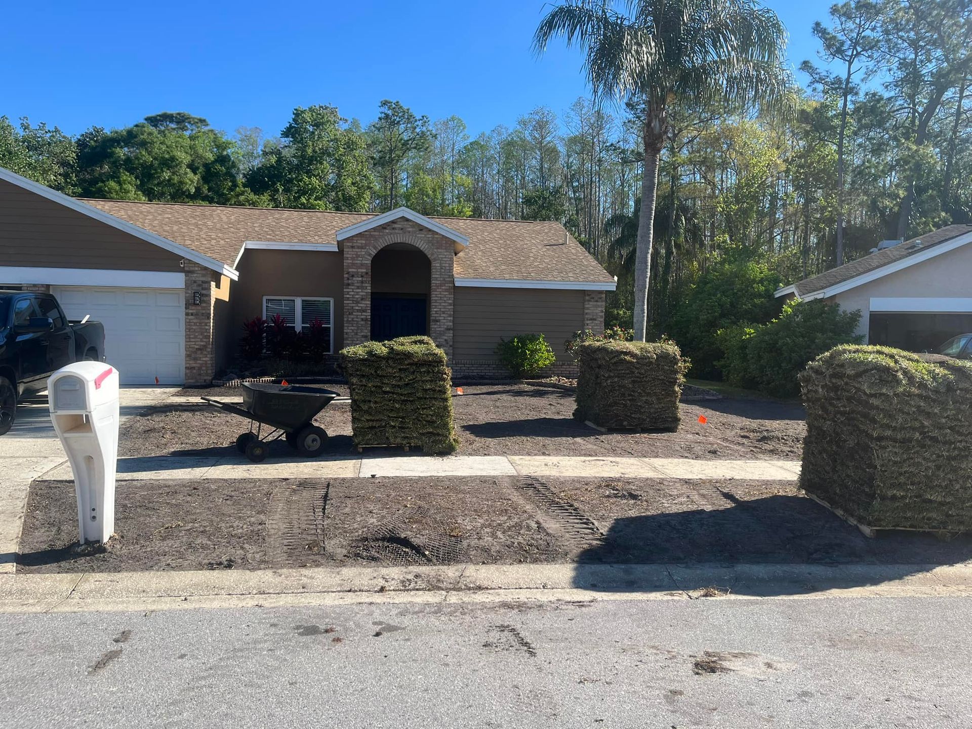 A suburban house front yard with a driveway, a mailbox, and several stacks of sod pallets ready for landscaping.