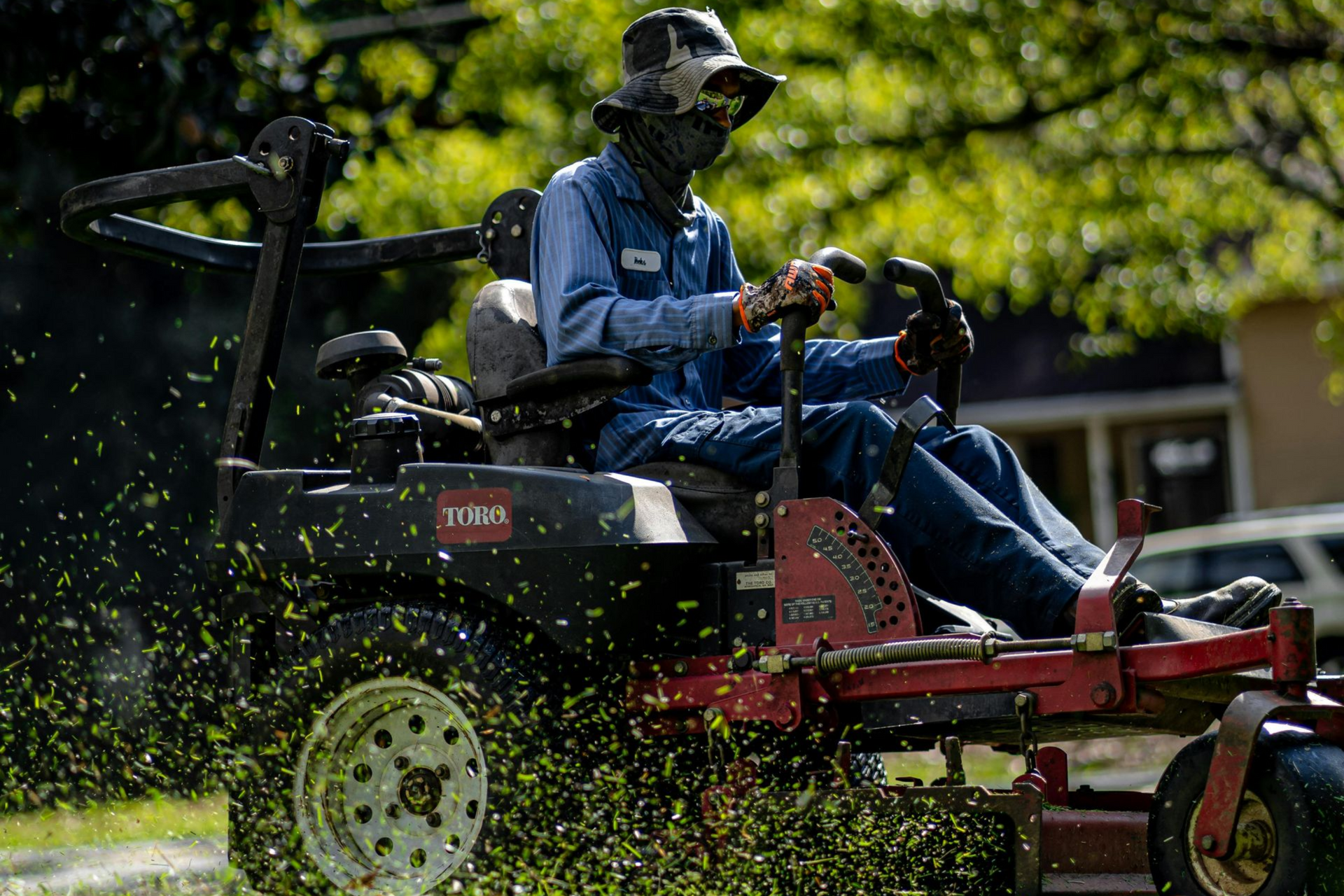 A person wearing protective gear and a hat operates a red zero-turn lawn mower, spraying grass clippings while working.