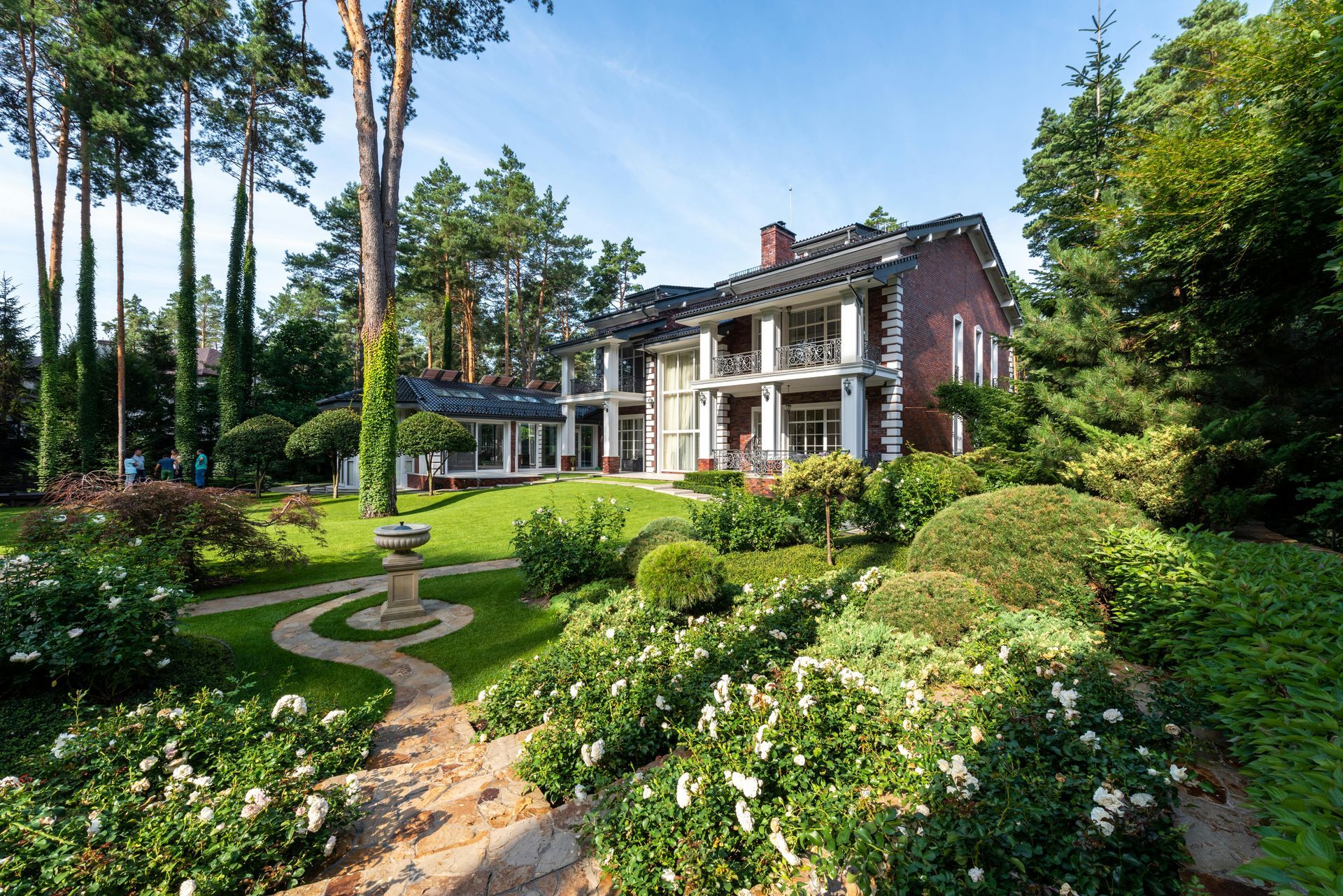 A multi-story brick house with white columns sits amidst a lush green garden with white flowers and tall trees.