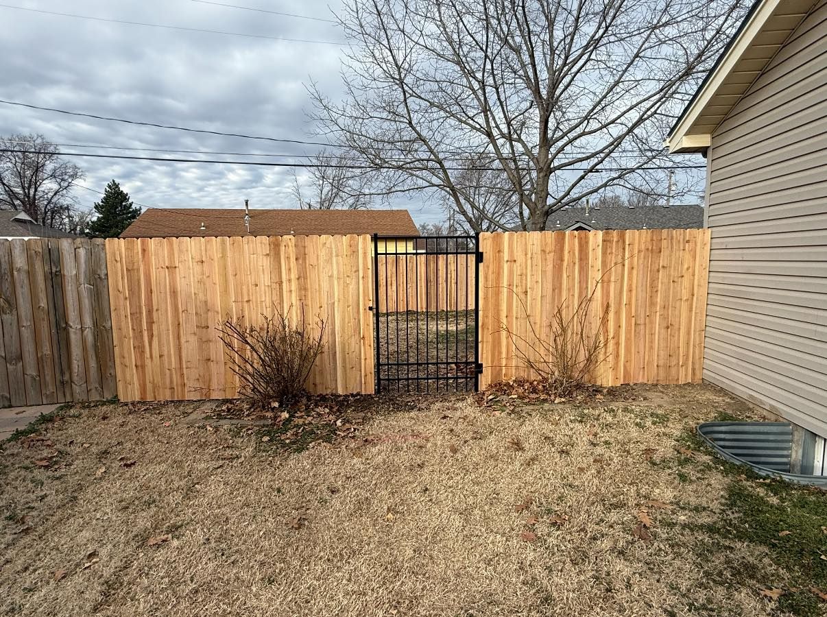A wooden fence with a central black metal gate stands in a backyard under a cloudy sky.