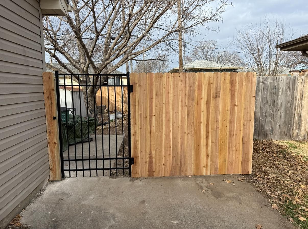 A metal gate installed between a house wall and a new wooden privacy fence on a concrete driveway.