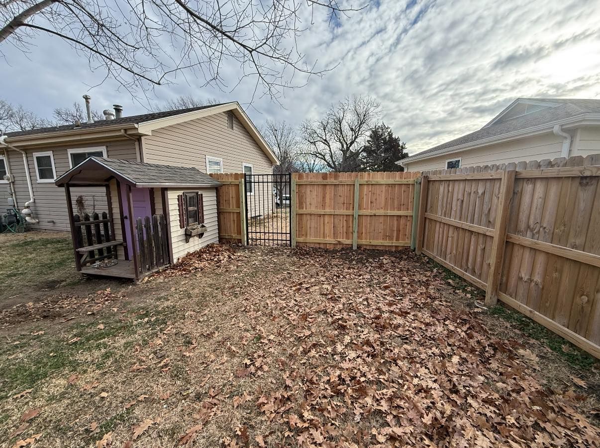 A fenced-in backyard covered in brown leaves, featuring a small wooden playhouse next to a wooden fence and metal gate.