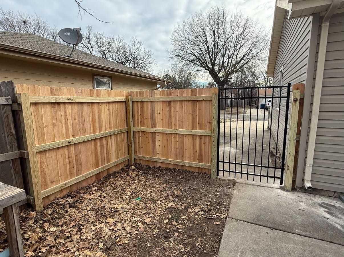 A wooden fence connects to a metal gate next to a house, bordering a dirt yard covered in fallen leaves.