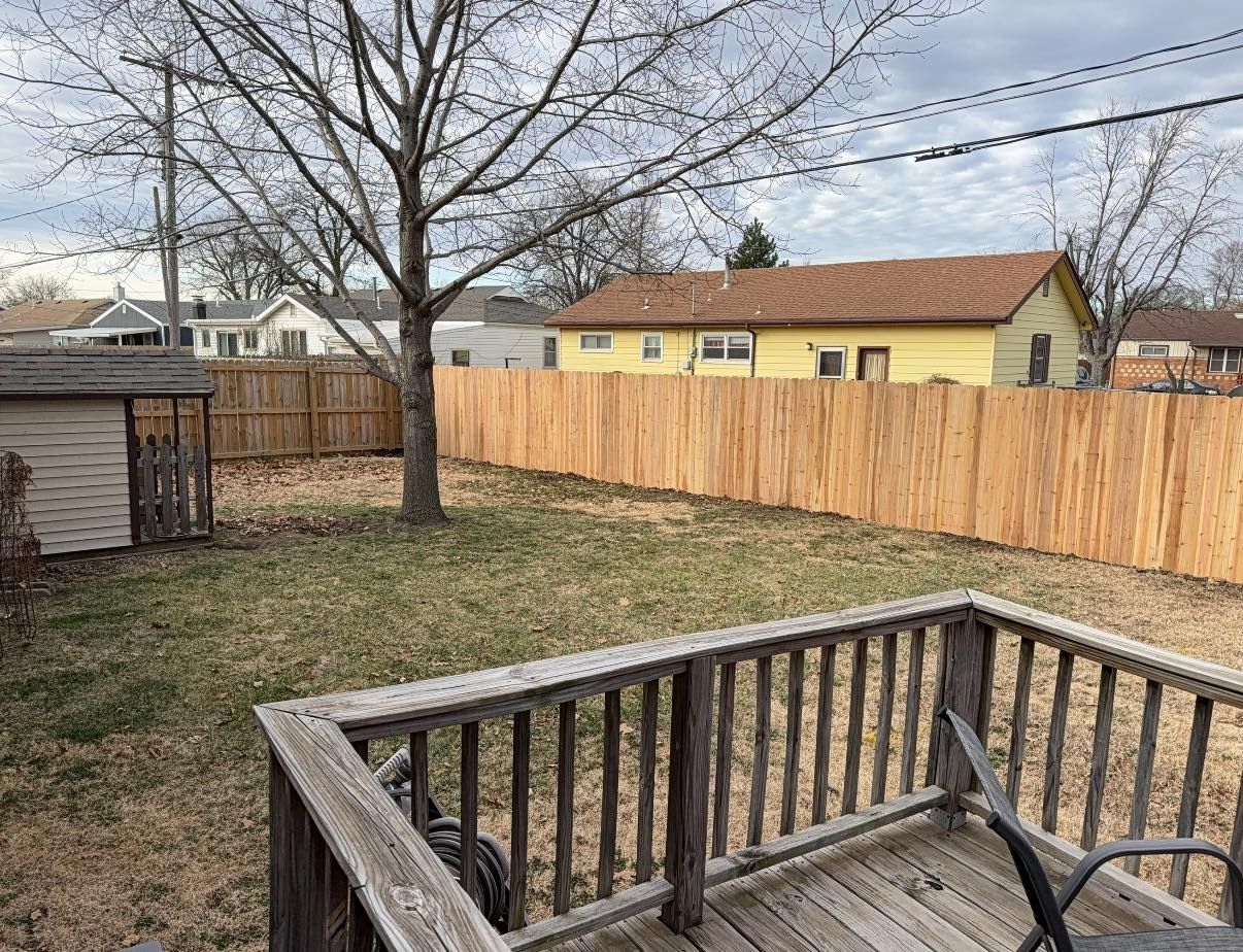 A wooden deck overlooks a grassy backyard with a tall wooden privacy fence and a yellow house in the background.