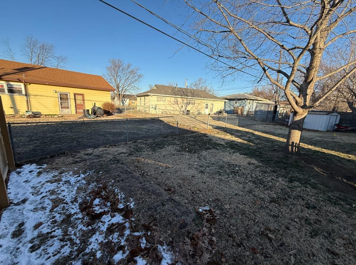 A backyard scene with patches of snow on the ground, a yellow house on the left, and a bare tree against a blue sky.