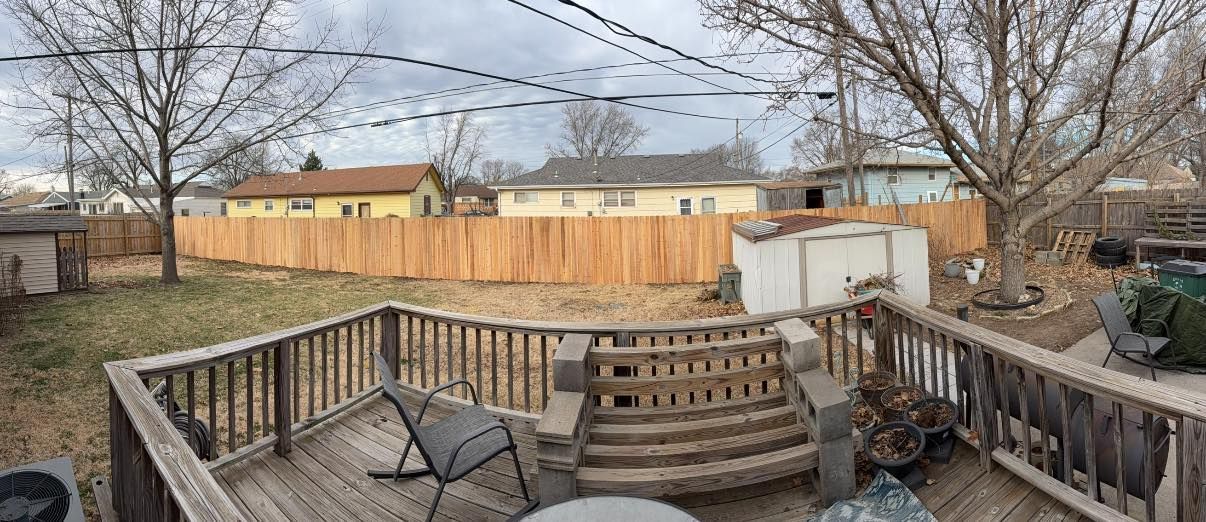 A wooden deck with patio furniture overlooks a backyard featuring a tall wooden fence and nearby residential houses.