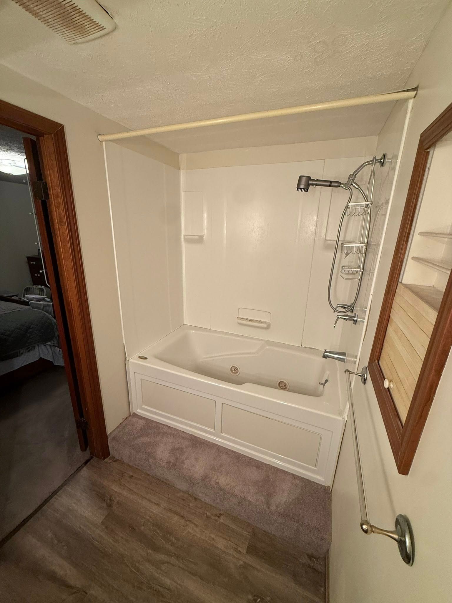 A white jetted bathtub with a handheld shower head in a tiled alcove, next to a doorway and a wood-framed shelving unit.