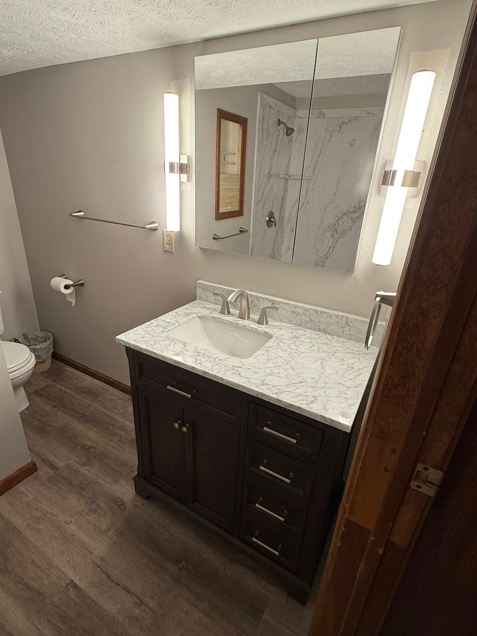 A modern bathroom featuring a dark wood vanity with a white marble countertop, a wall mirror, and vertical sconce lights.
