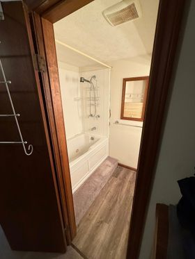 A doorway looking into a bathroom featuring a white tub-shower combo, wood-look flooring, and a small wooden mirror.