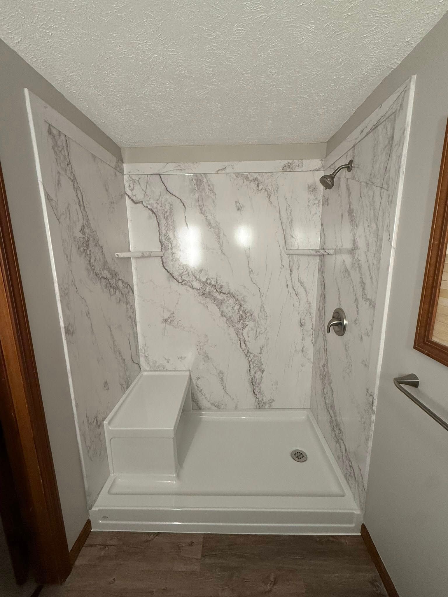 A walk-in shower with white marble-patterned walls, a built-in corner seat, chrome fixtures, and a white shower pan.