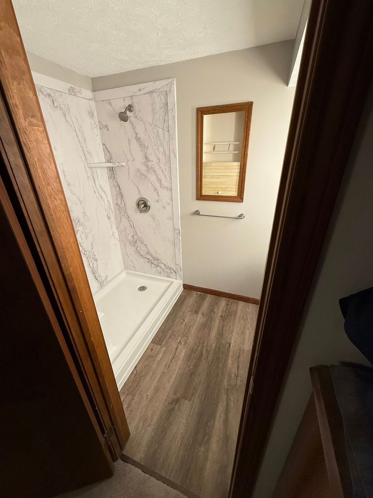 A bathroom with light gray walls, wood-look flooring, a walk-in shower with white marble-patterned walls, and a wall mirror.