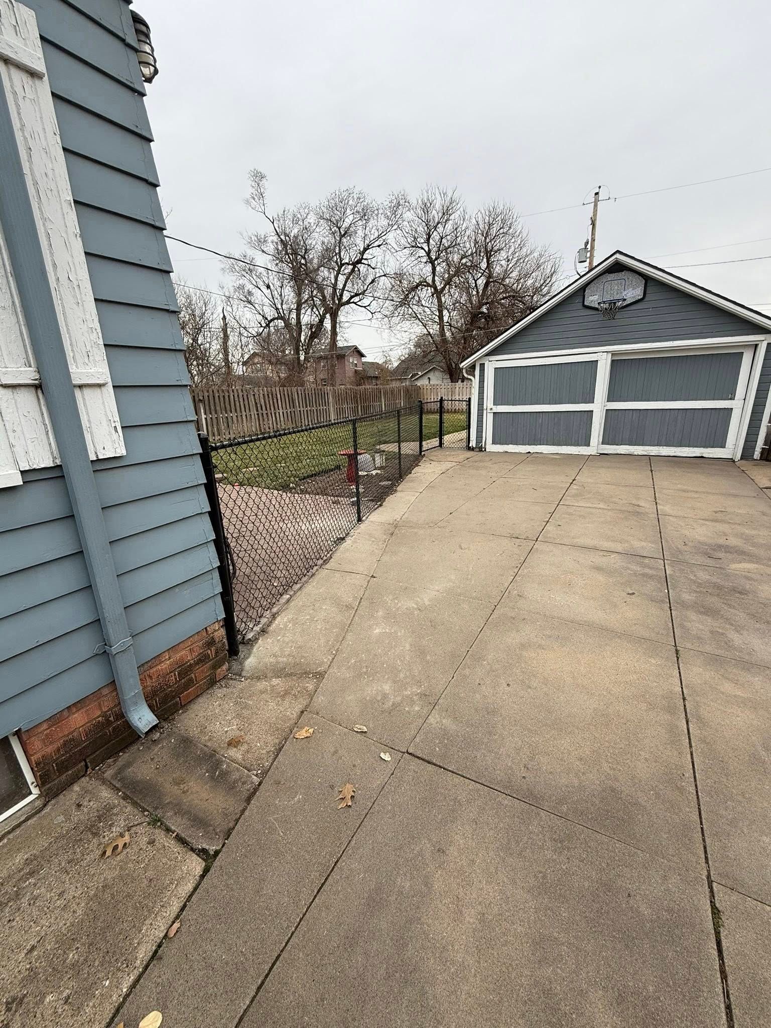 A gray-sided house exterior with a concrete driveway leading to a detached garage under an overcast sky.