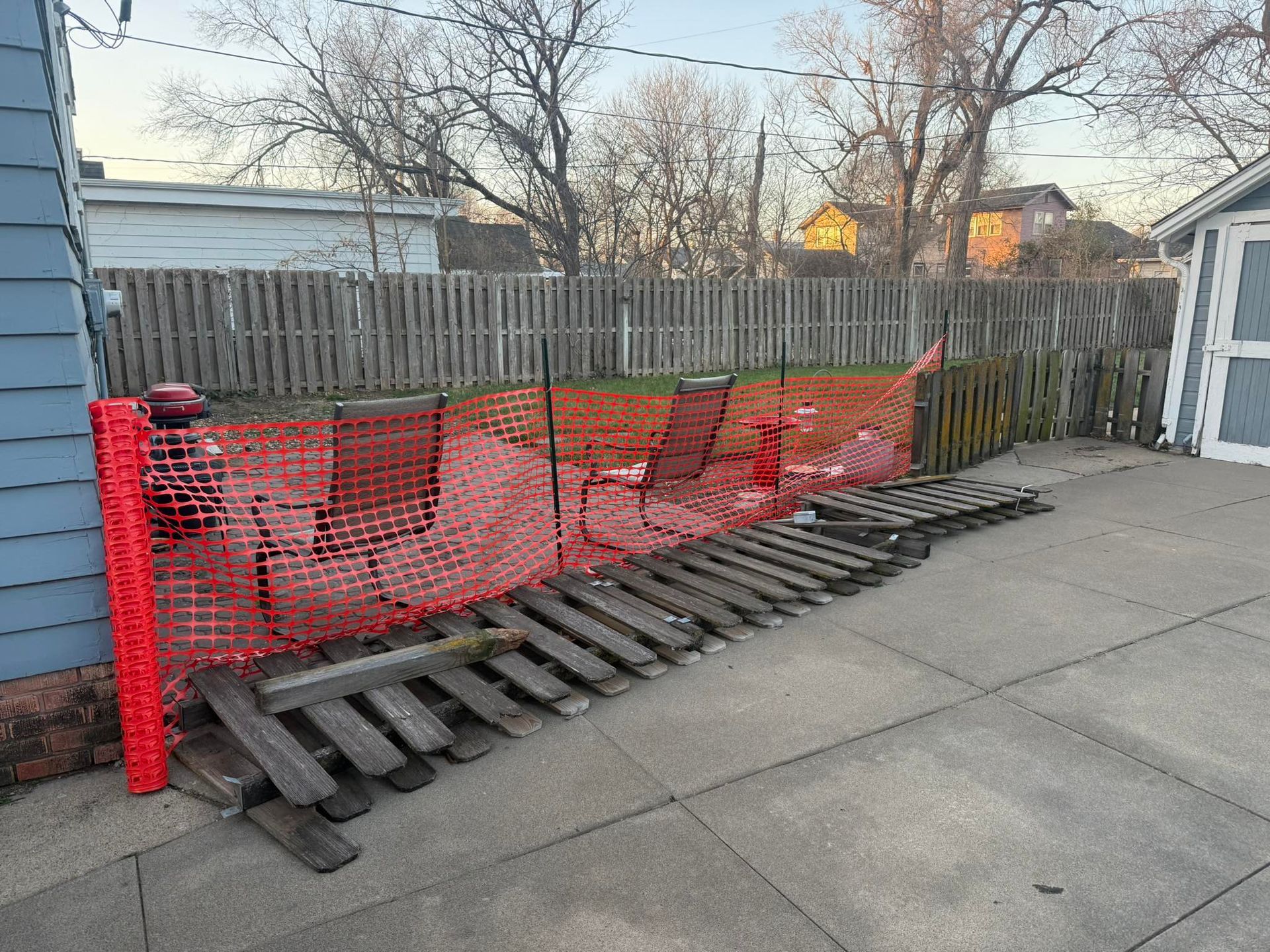 A fallen wooden fence panel lies on a concrete patio in front of a temporary orange plastic construction fence.