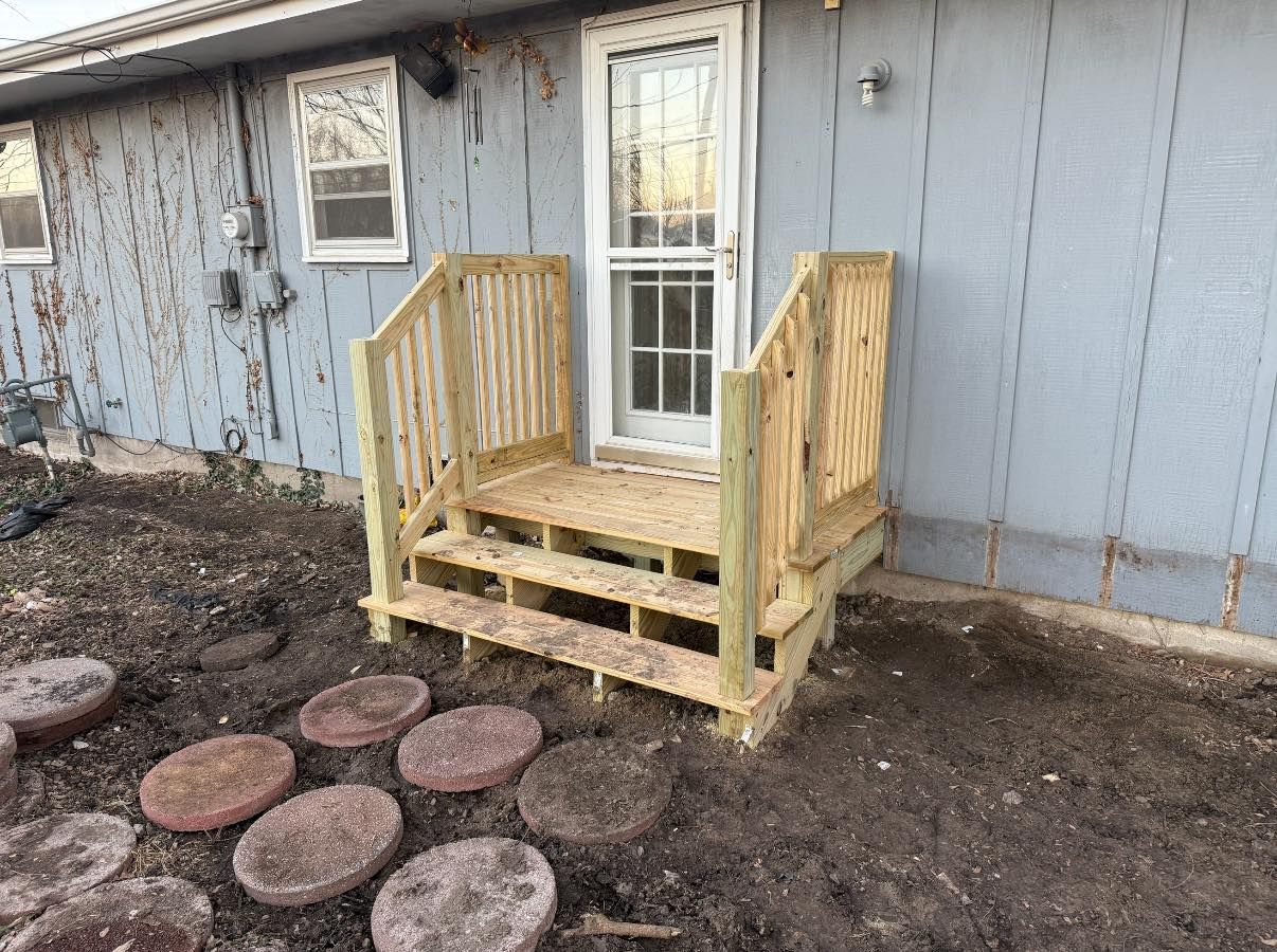 A newly built wooden deck with two steps attached to the back entrance of a gray house, set over a dirt yard.