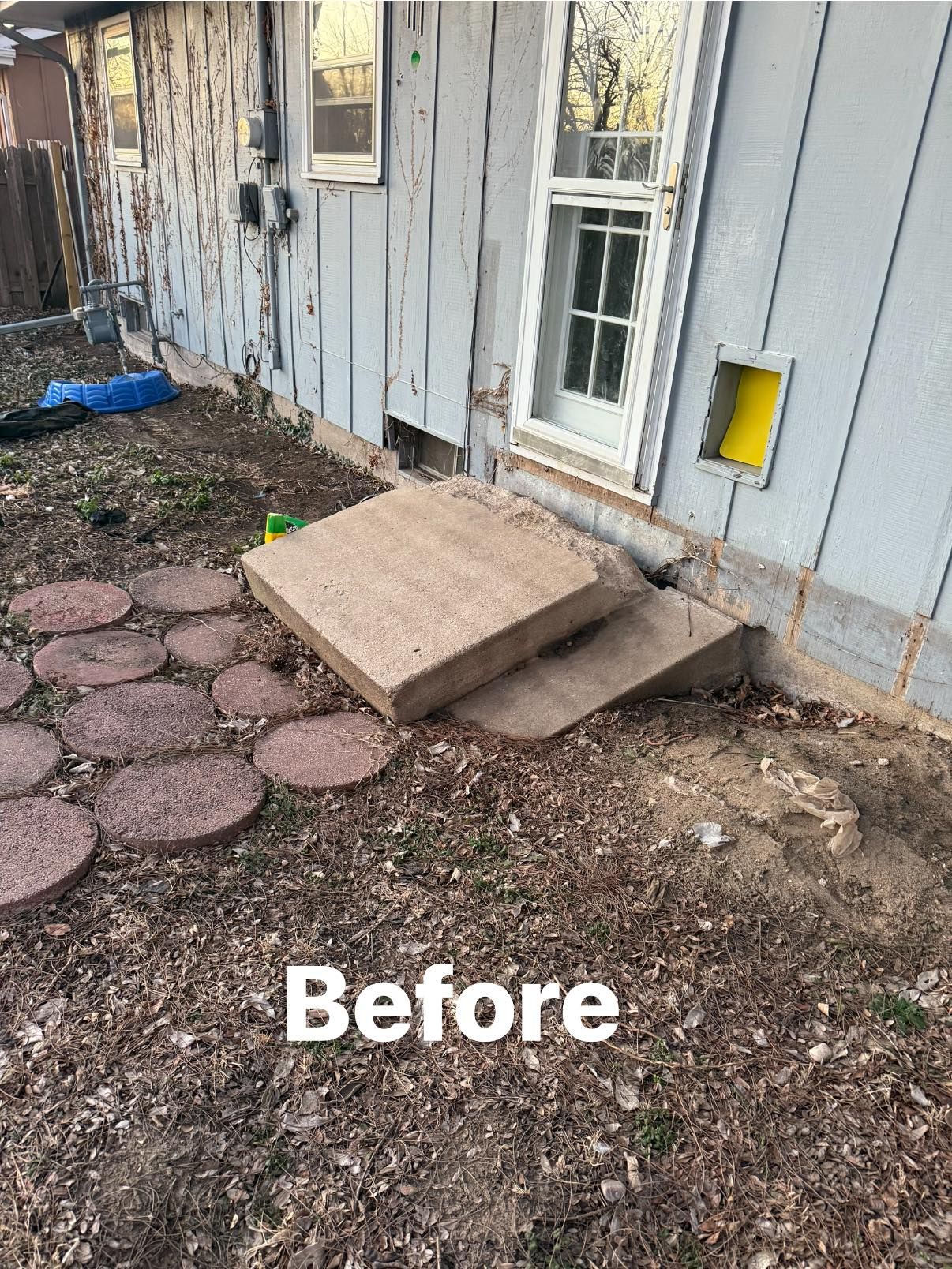 A dilapidated concrete slab landing tilted away from the side door of a house with grey siding and a yellow pet door.