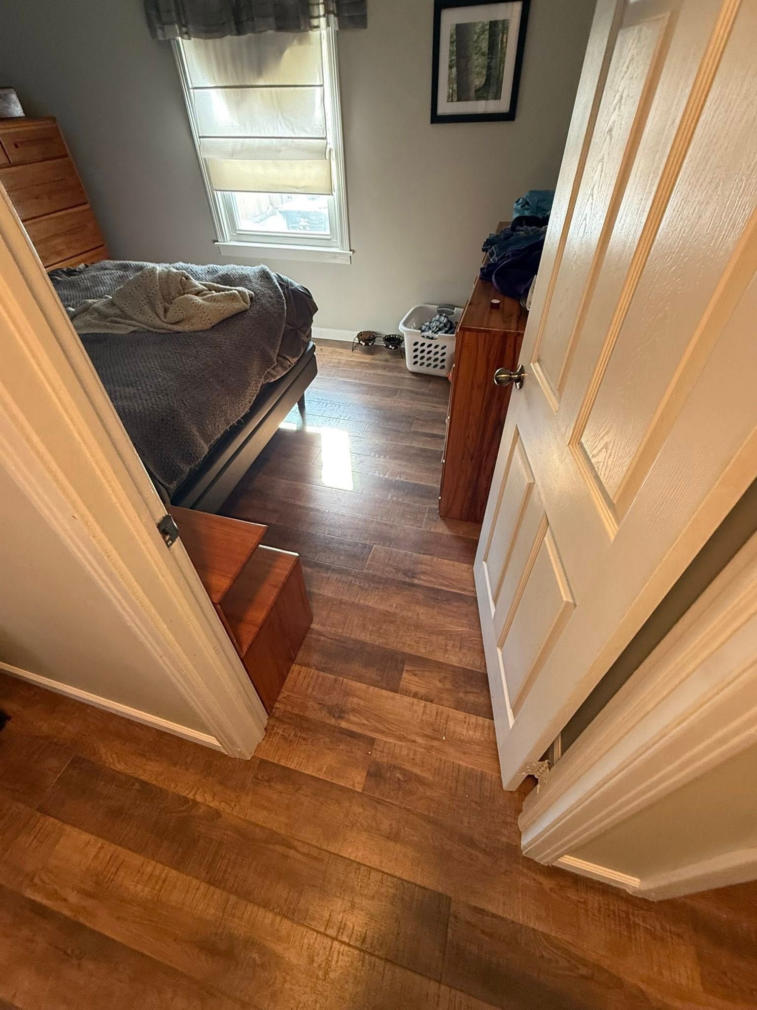 A view from a doorway into a small bedroom with a bed, a wood dresser, a window with blinds, and wood-patterned flooring.