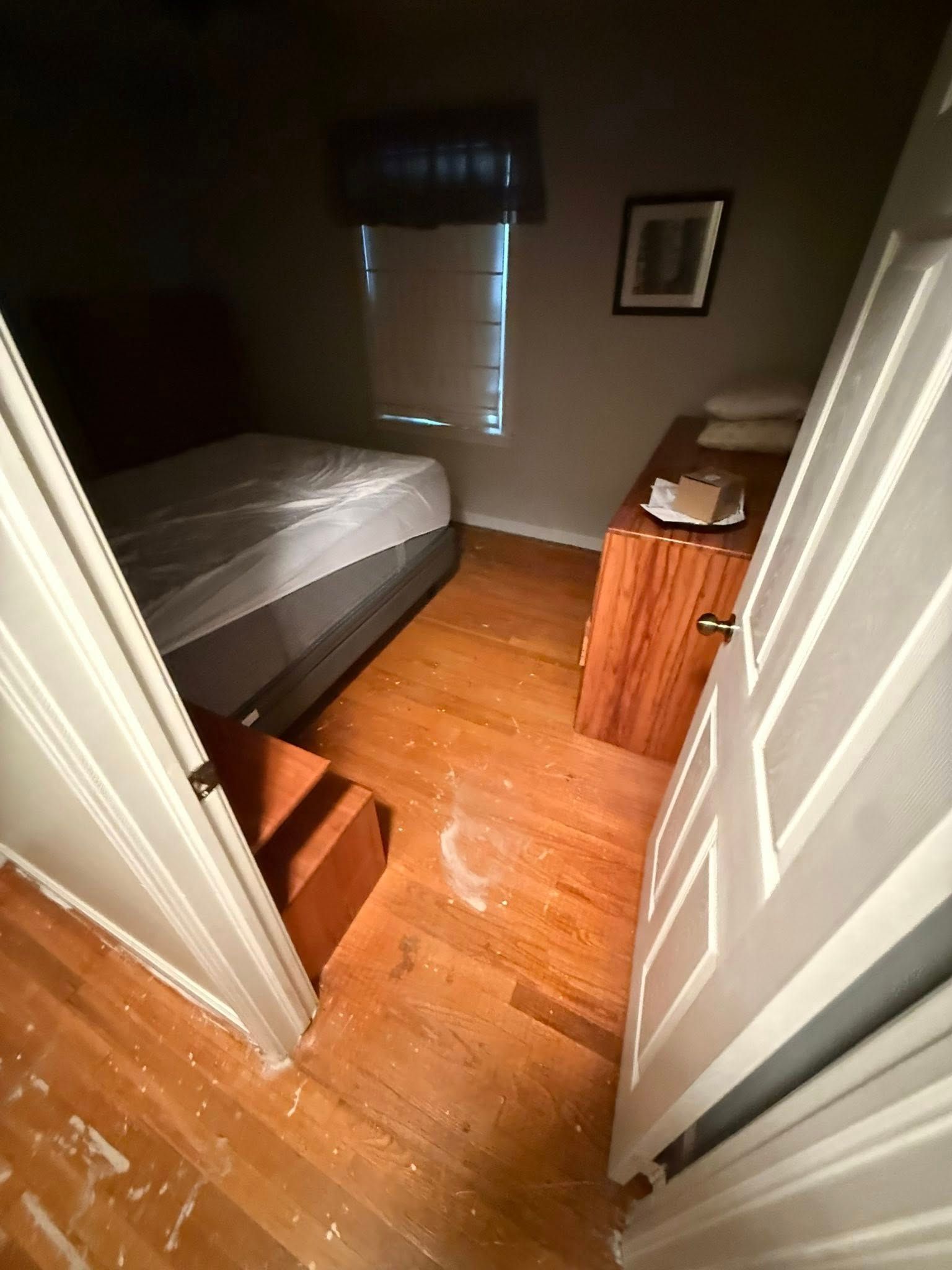 A bedroom view from a doorway, featuring a bed with white bedding, a wooden dresser, and light-colored hardwood flooring.