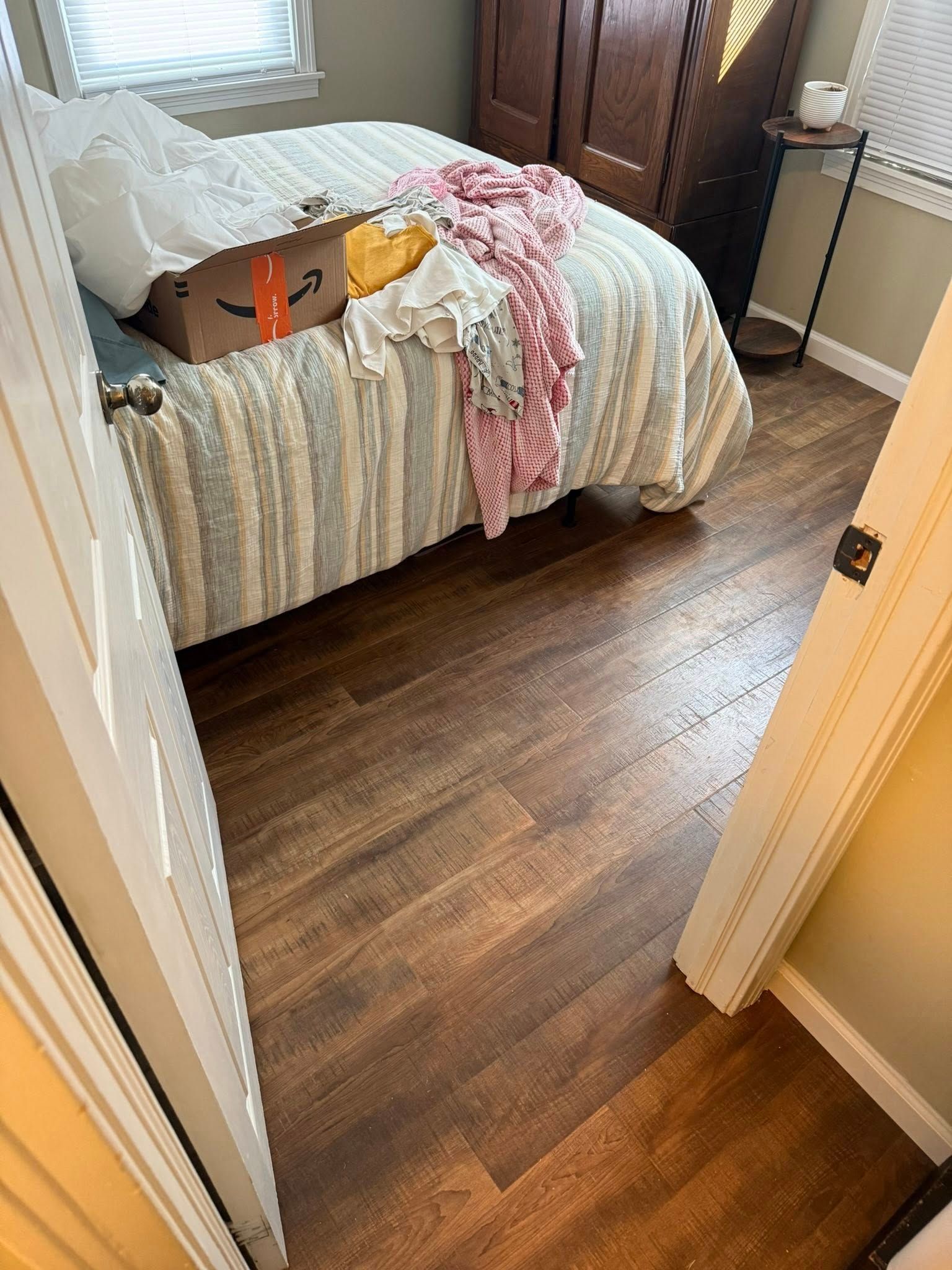 A bedroom view from a doorway, featuring a bed with a striped blanket, a cardboard box, and clothes on wood-look flooring.