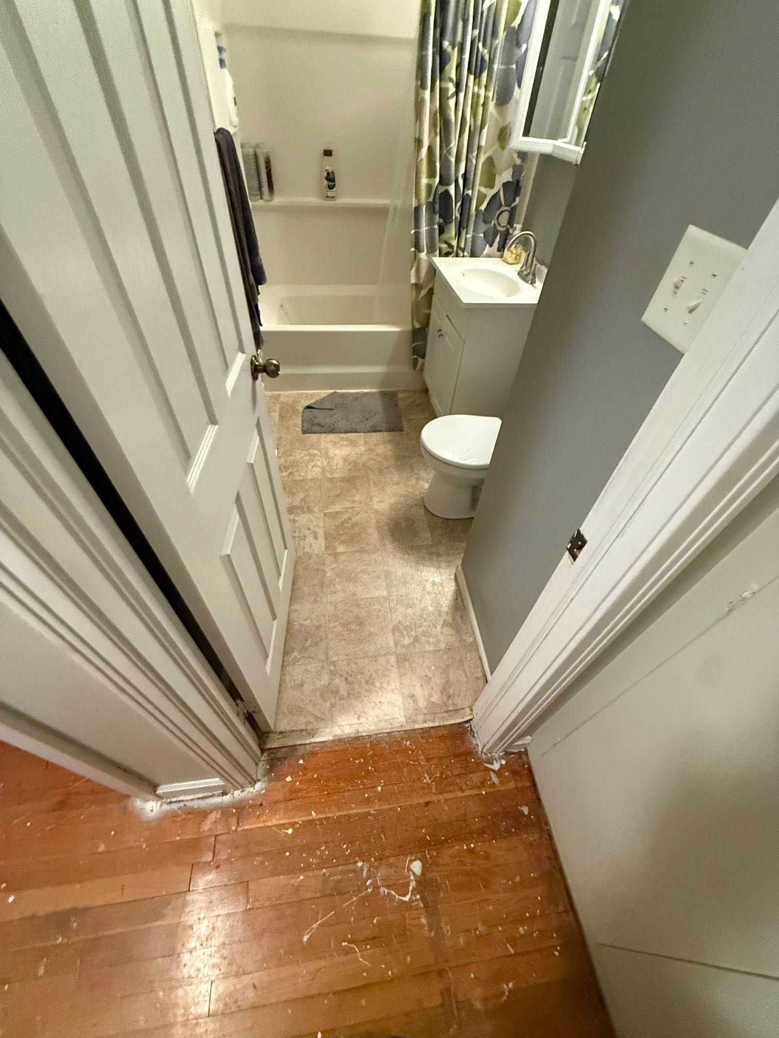 A view through an open doorway into a small bathroom with a tub, toilet, and vanity, leading to hardwood flooring.