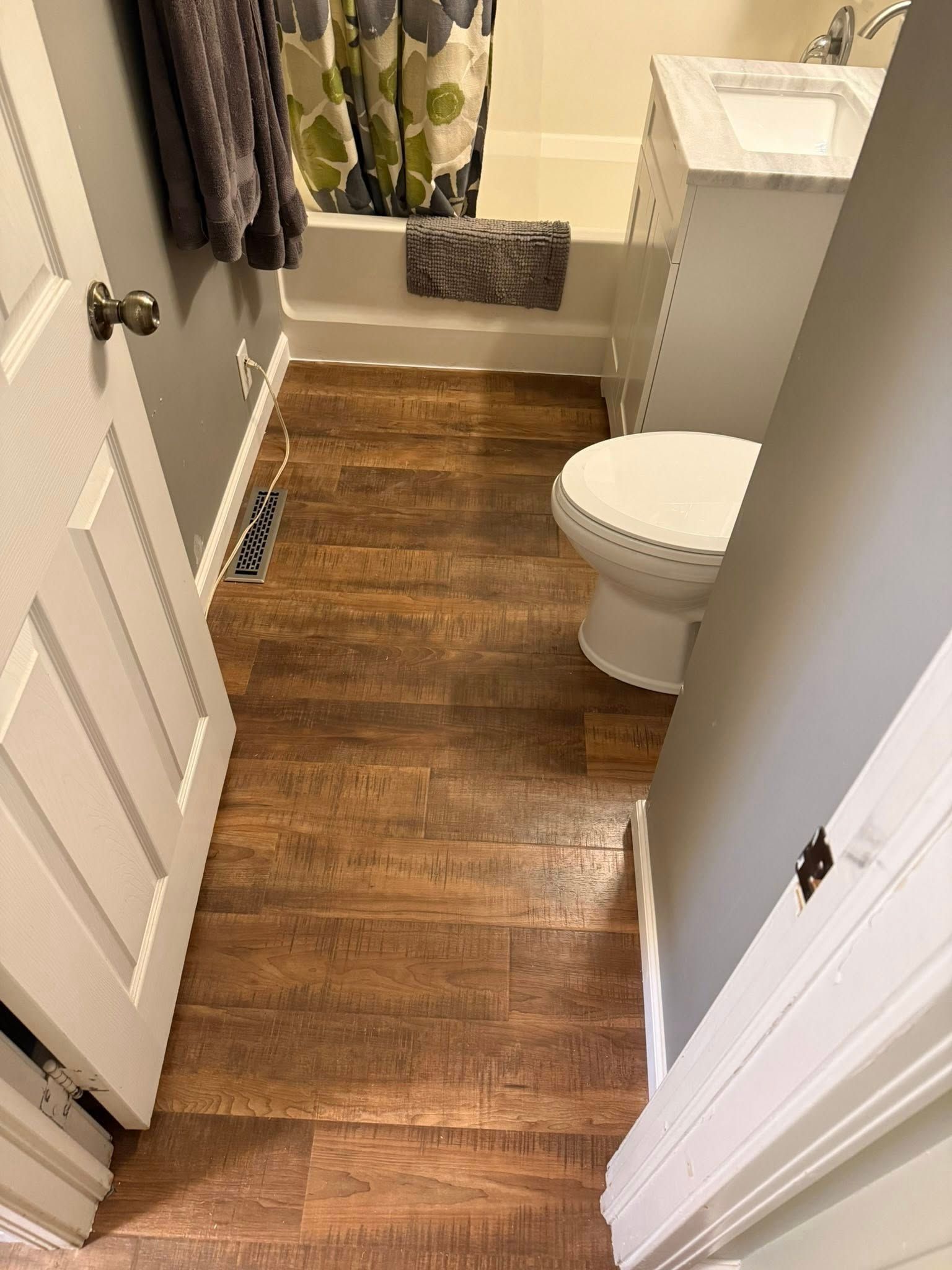A narrow bathroom with wood-look flooring, a white vanity, a toilet, and a bathtub with a patterned shower curtain.