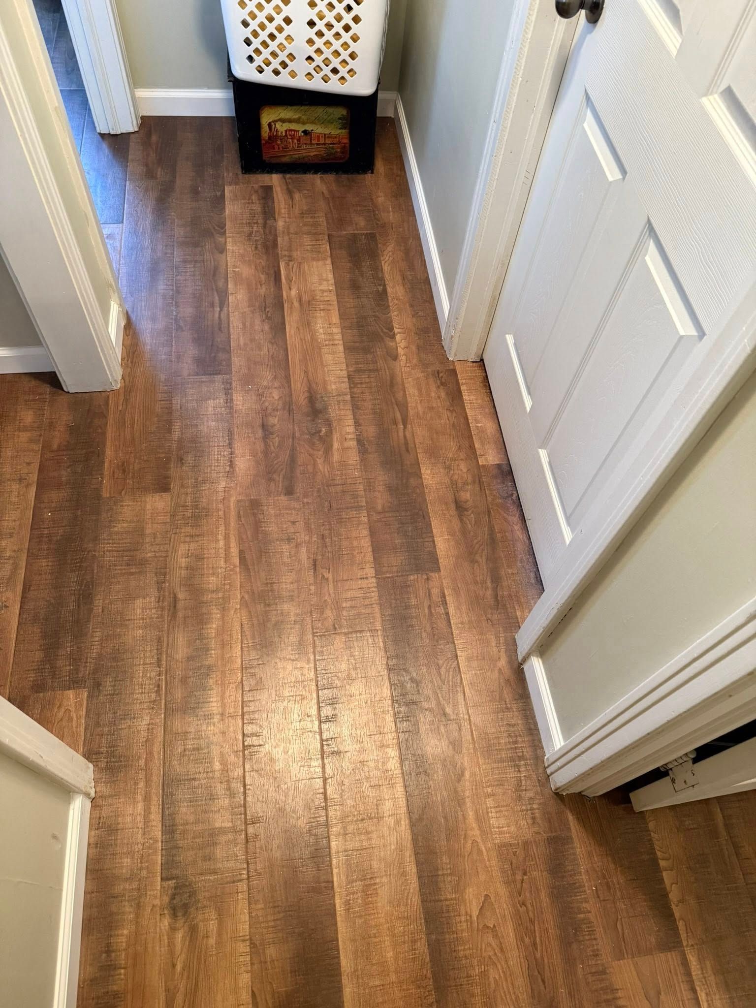 A high-angle view of a narrow hallway with rustic, dark brown wood-look flooring, white trim, and a closed white door.