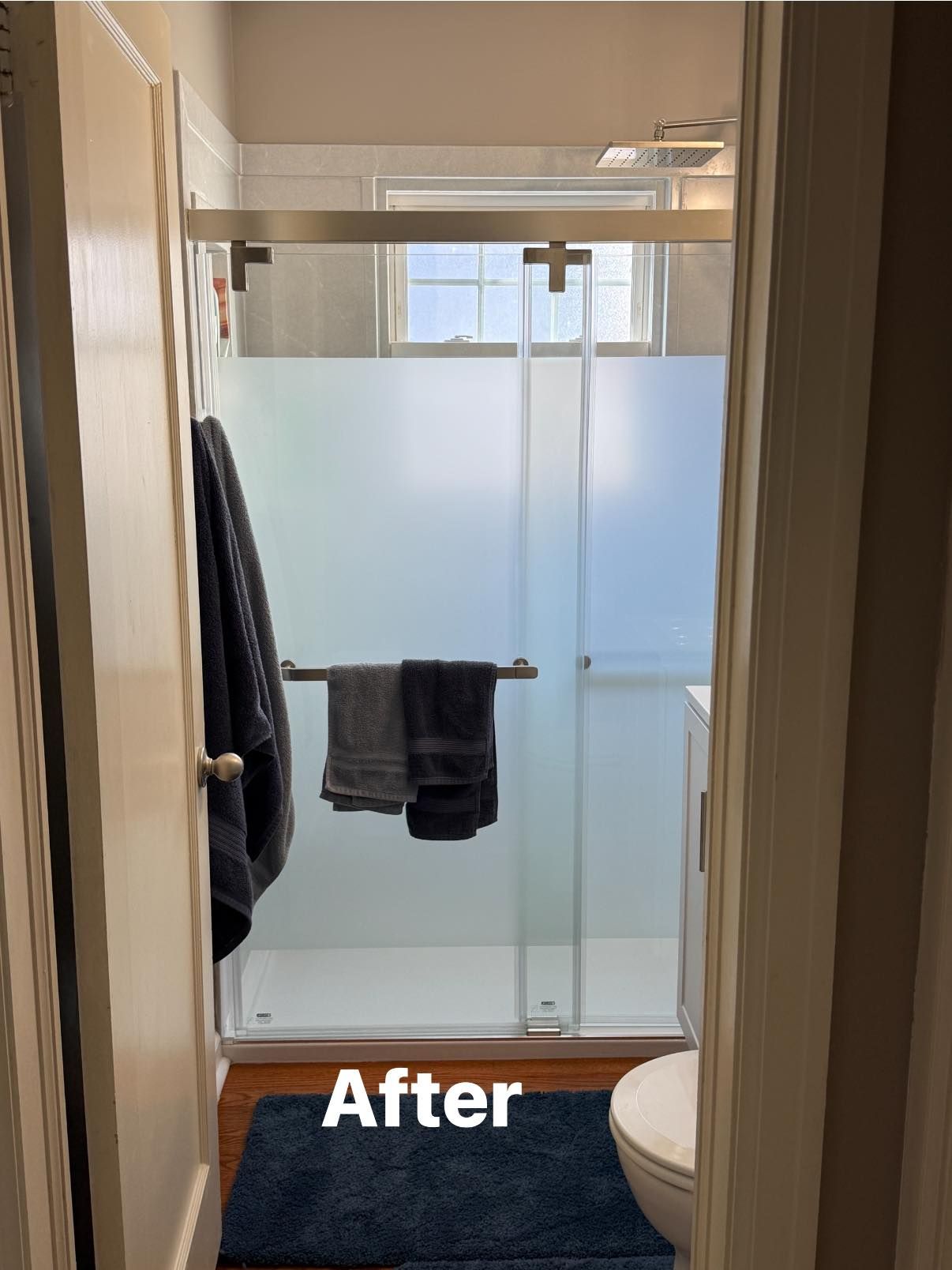 A bathroom with a new walk-in shower featuring frosted glass sliding doors, a dark blue rug, and hanging towels.