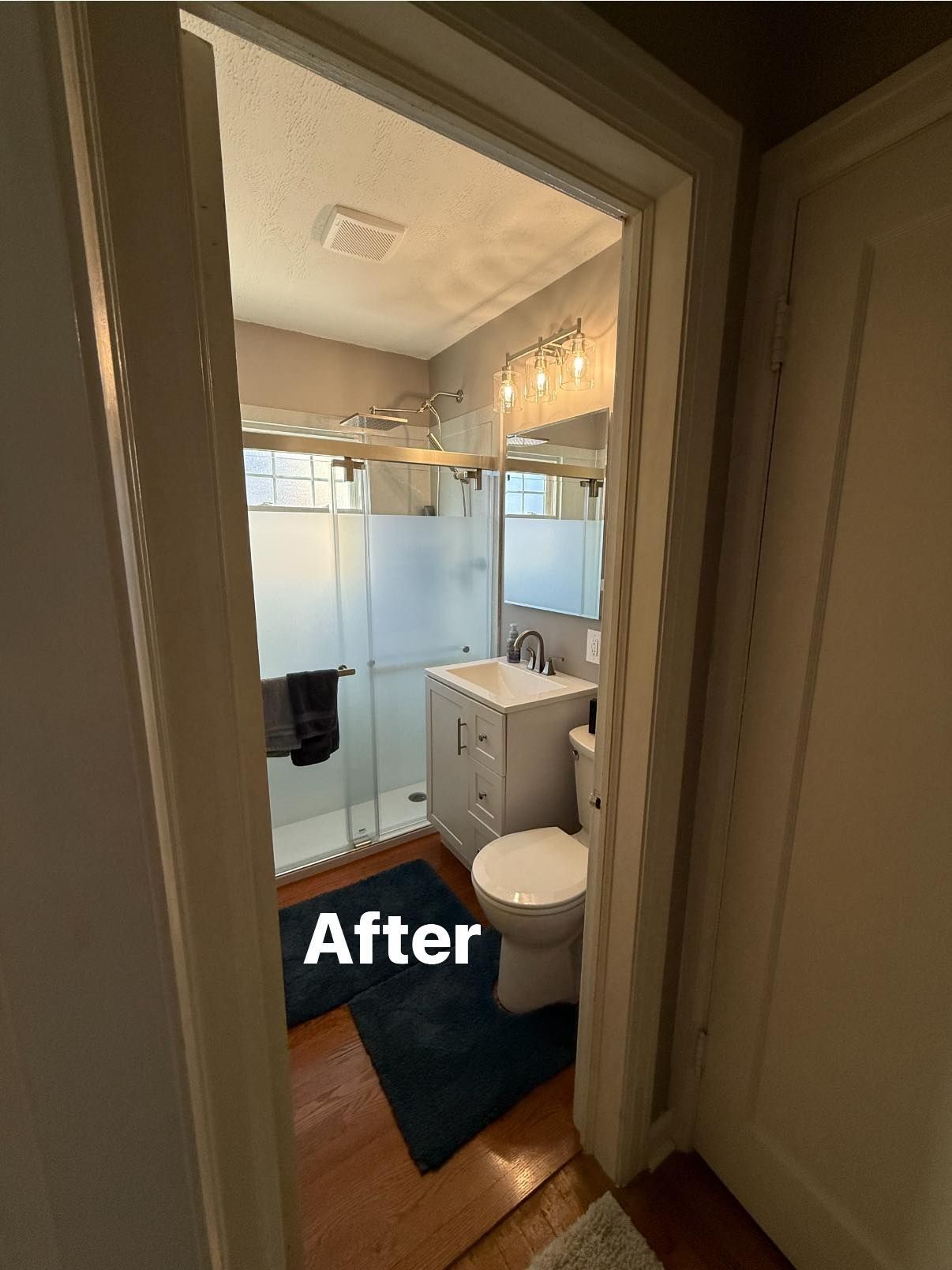 View through a doorway into a renovated bathroom featuring a white vanity, a glass shower, and a blue rug on wood floors.