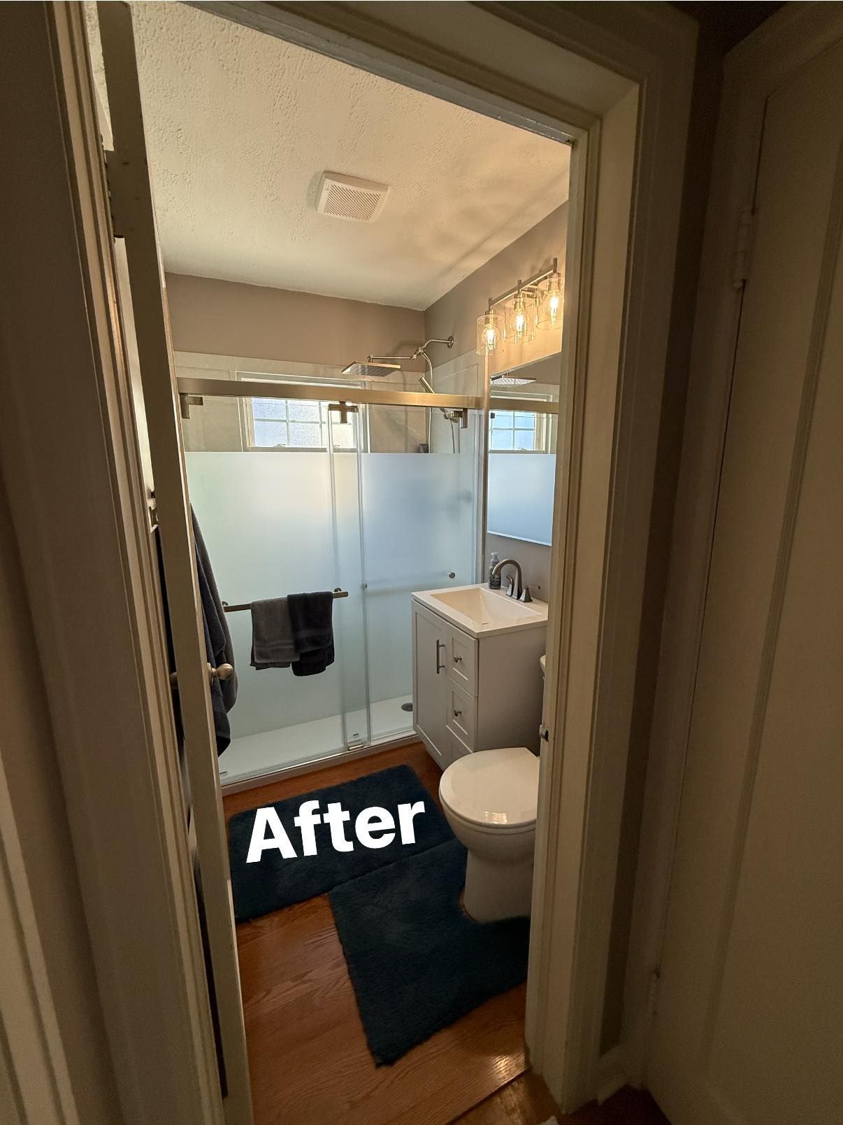 A bathroom with a frosted glass shower door, a white vanity, a toilet, and dark blue floor mats, viewed from the doorway.