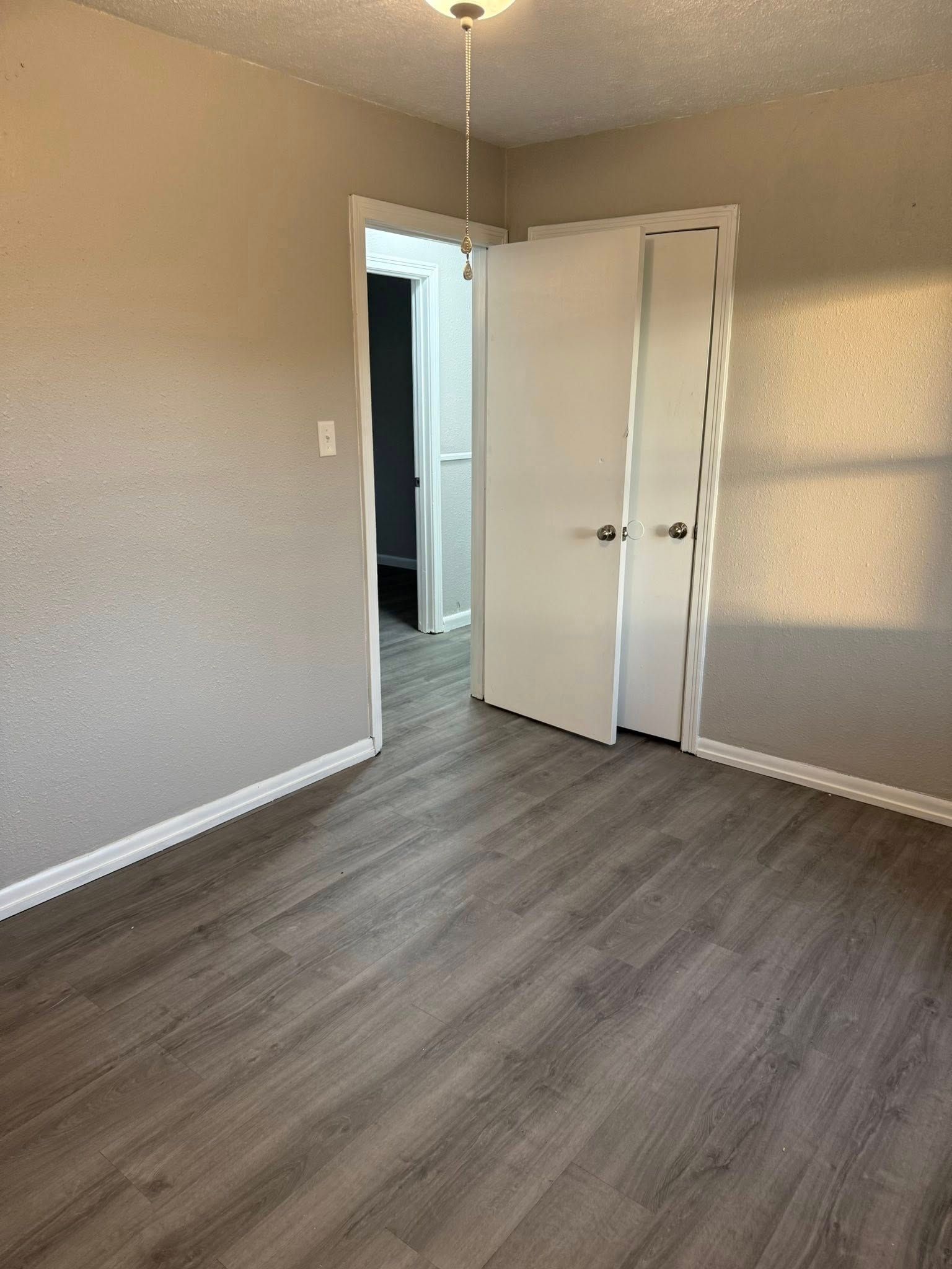An empty bedroom with gray wood-look flooring, light-colored walls, and an open white door leading to another room.