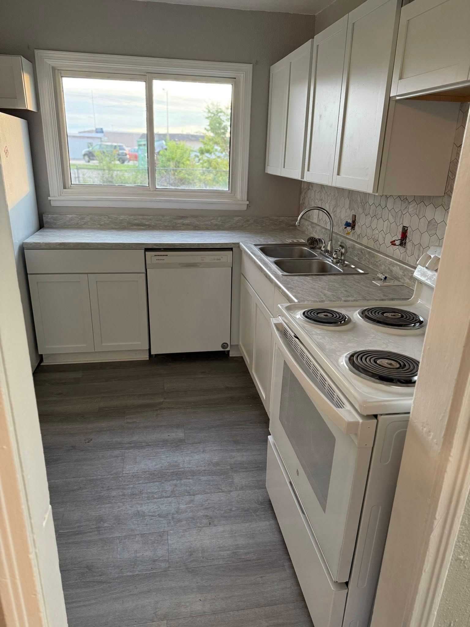 A narrow kitchen with white cabinets, light countertops, a white oven, a dishwasher, and gray wood-look flooring.