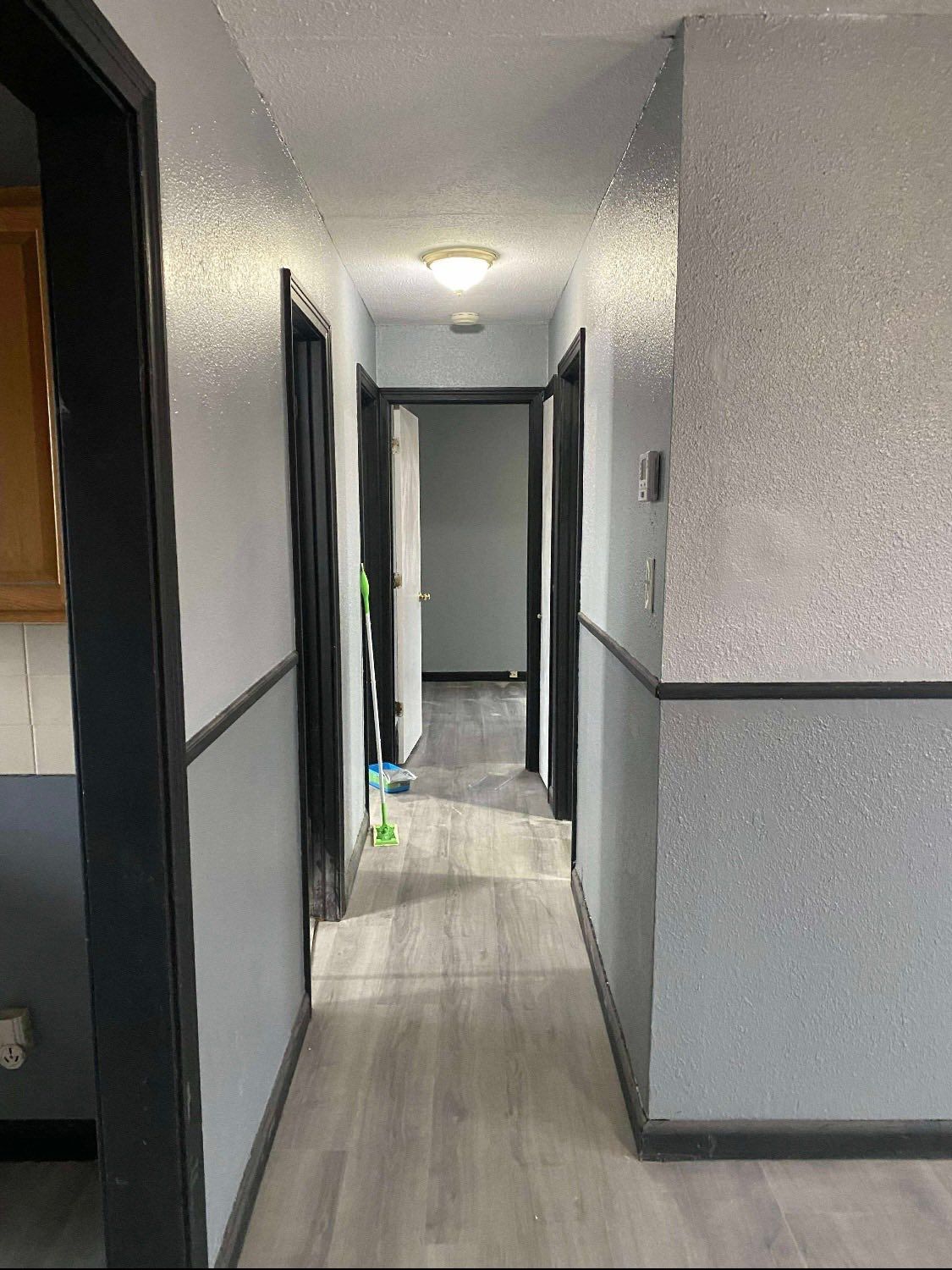 A hallway with light gray textured walls, a dark gray wainscoting, black door frames, and light wood-look vinyl flooring.