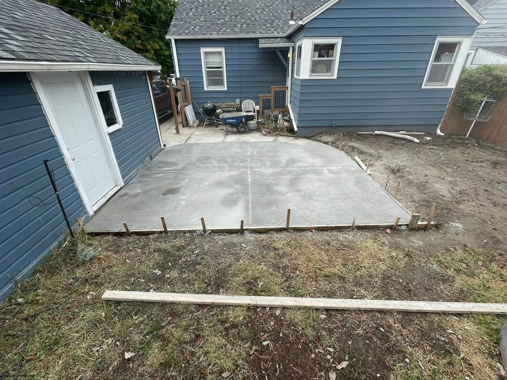 A newly poured concrete patio slab with wooden formwork surrounds, situated in a backyard between a house and garage.