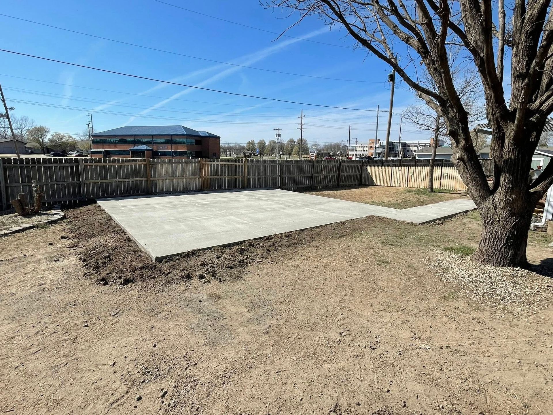 A newly poured concrete rectangular patio sits in a backyard near a large tree, bordered by a wooden fence under blue sky.