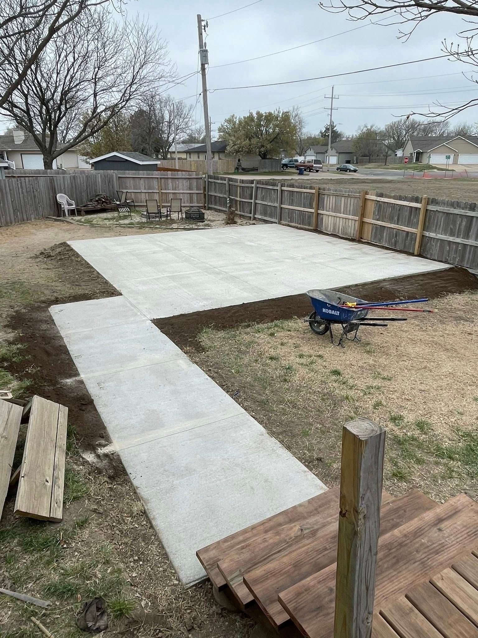 A new concrete patio and walkway extend from wooden steps into a fenced backyard.