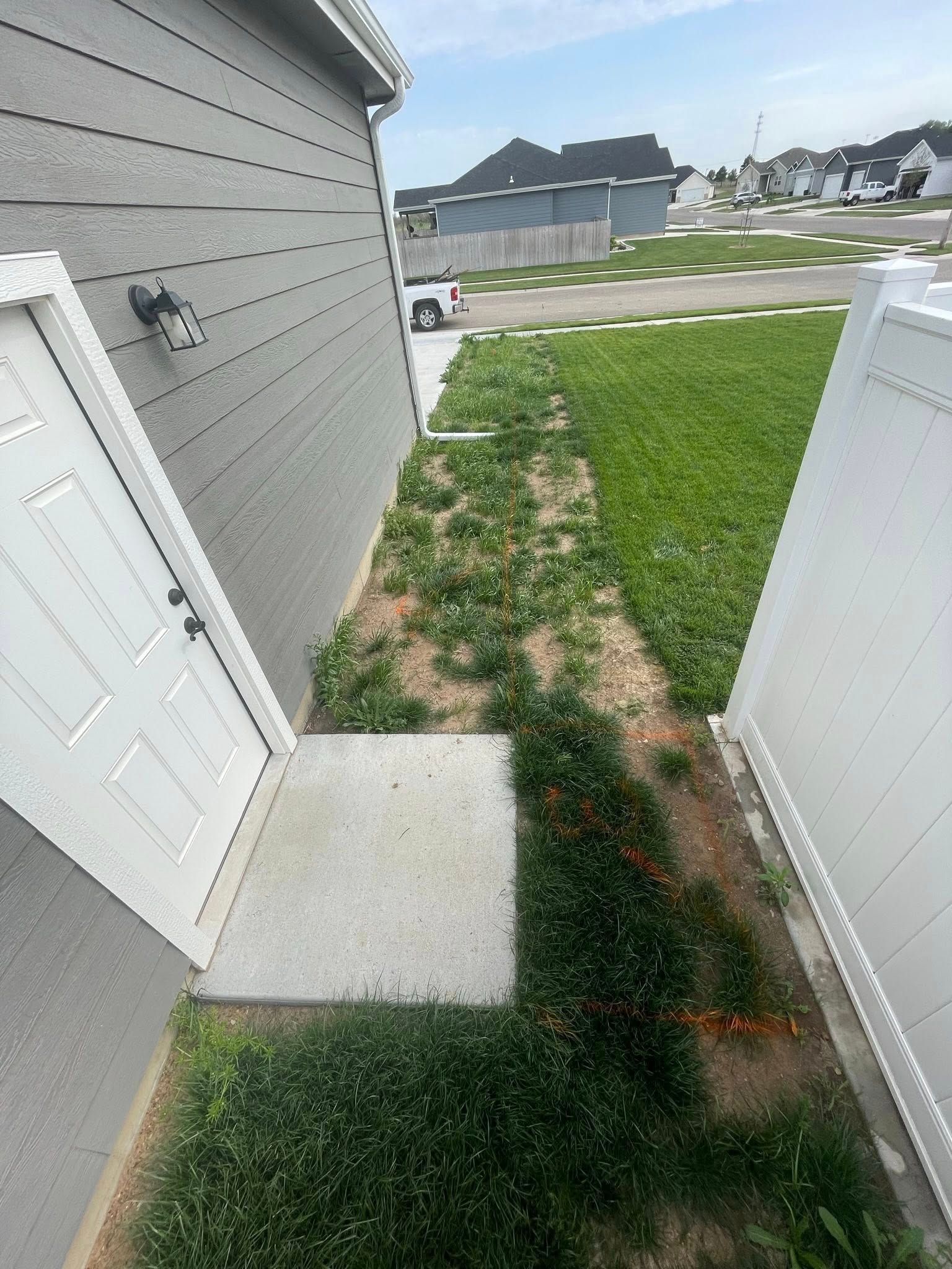 A narrow outdoor side yard between a gray house wall and a white fence, featuring a concrete pad and patchy grass.