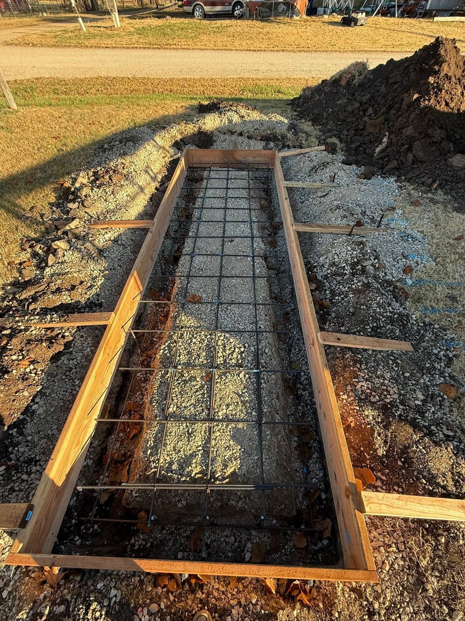 A wooden form frame set on gravel for a concrete pour, featuring a metal wire grid interior, outdoors at a construction site.