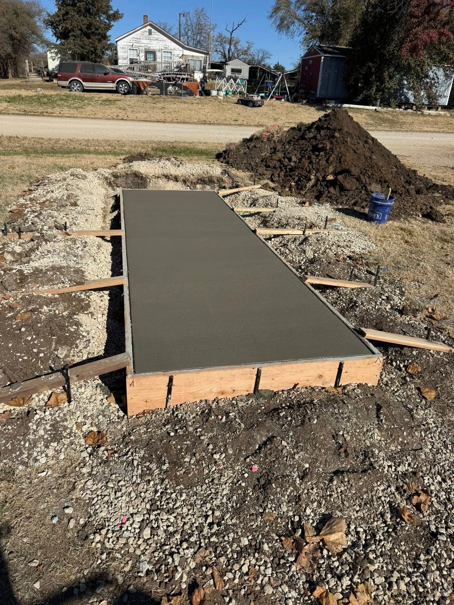 A fresh concrete walkway slab encased in wooden forms, set in a gravel-filled yard with a house visible in the background.