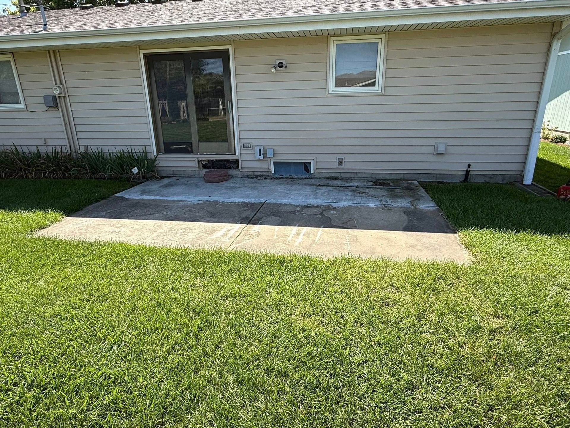 A small, weathered concrete patio sits against the beige siding of a house with a sliding glass door and one window.