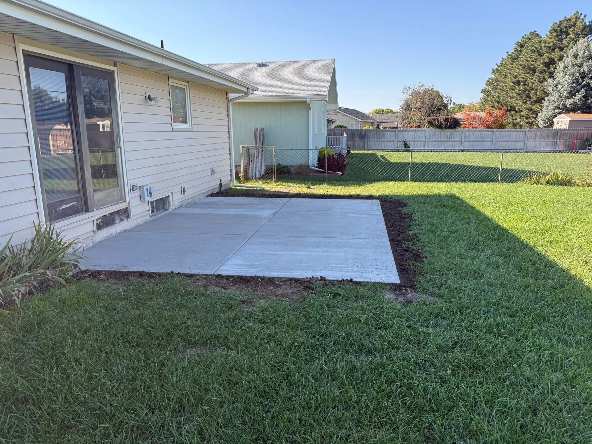 A rectangular concrete patio sits beside a light-colored house in a sunny backyard with green grass and a wooden fence.