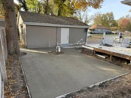 A newly poured concrete patio slab outside a residential garage next to a wooden deck on a sunny autumn day.