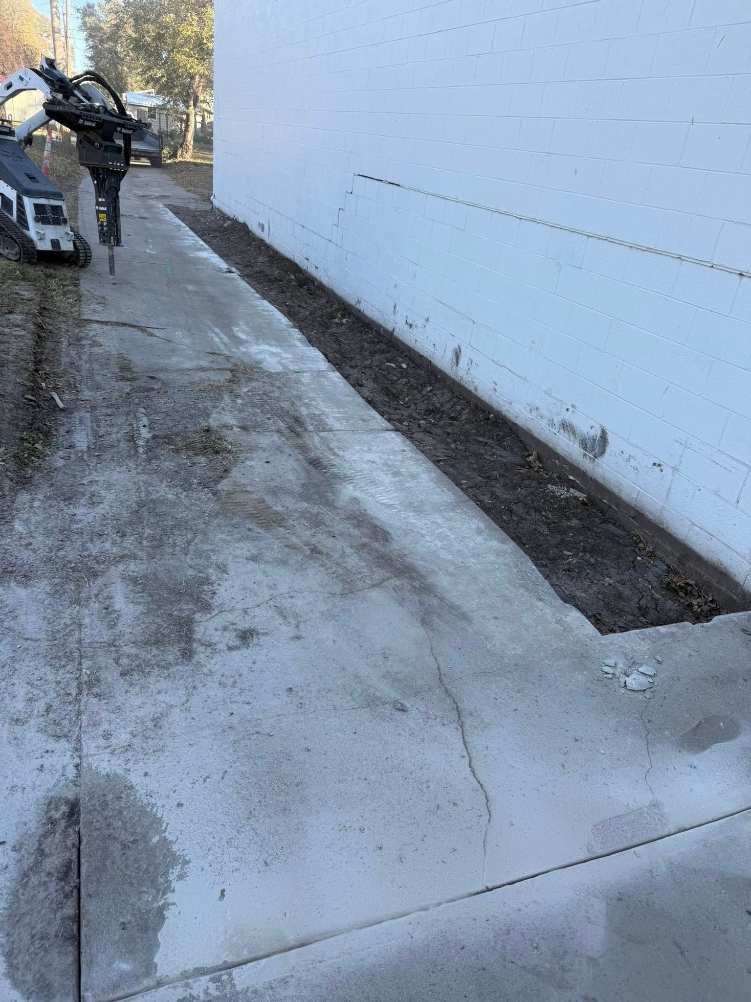 Construction machinery sits near a building exterior with a trench dug along the concrete wall.