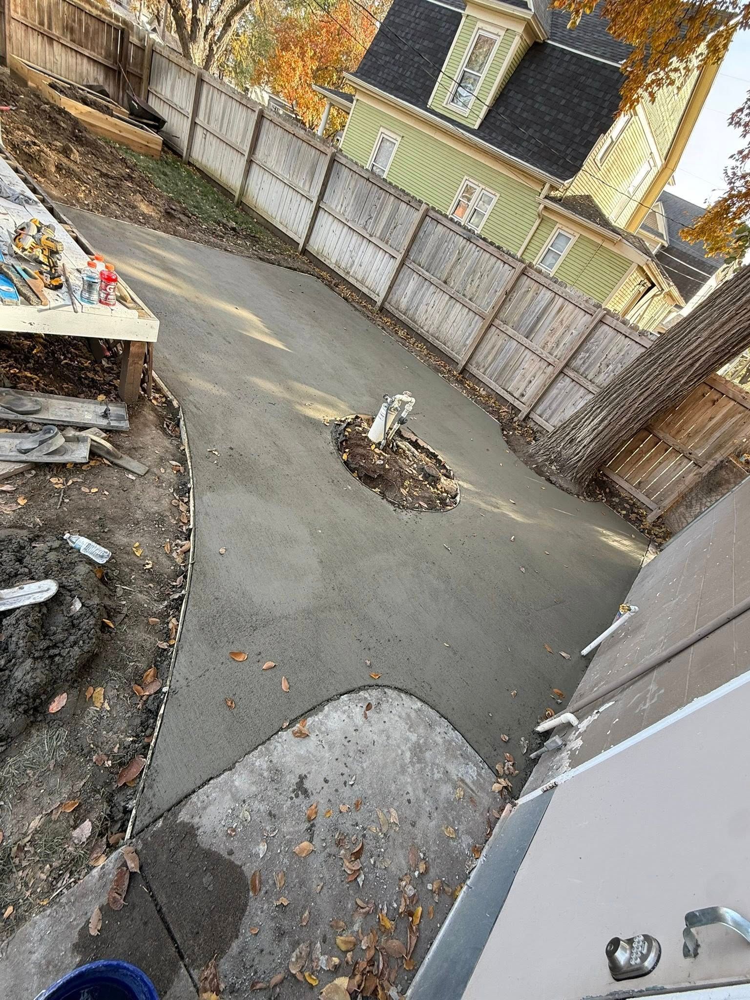 Freshly poured gray concrete patio in a backyard with a circular cutout around a post, bordered by a wooden fence.